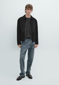 Schwarze Wildlederjacke mit Reißverschluss, zwei Vorderfächern, kombiniert mit einem dunklen Strickpullover, hellblauen Jeans und schwarzen Schuhen. Einheitlicher Hintergrund.