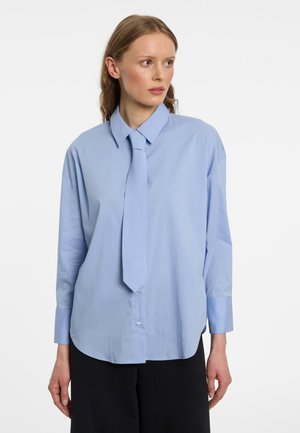 Femme aux cheveux châtain clair portant une chemise ample bleu clair à boutons et une cravate assortie, regardant vers sa gauche sur un fond uni.
