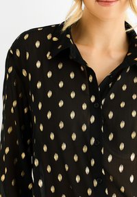 Black sheer blouse featuring gold oval patterns, a button-up front, and a collar. Material appears lightweight with a smooth texture.