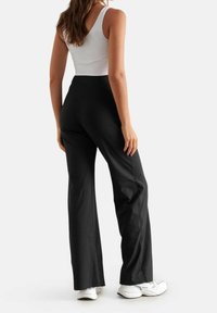 Pantalons noirs évasés avec une taille haute, fabriqués en tissu lisse. Associés à des baskets blanches, présentant un design simple et épuré.