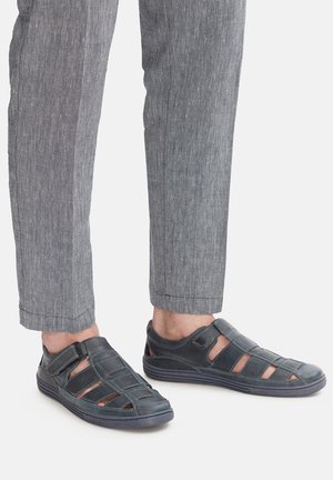 Sandali in pelle blu scuro con design aperto, caratterizzati da inserti, chiusura a cinturino e suola testurizzata; abbinati a pantaloni grigi e su misura.