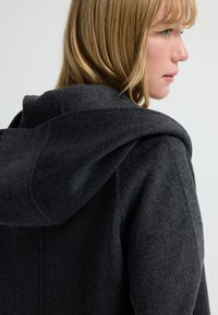 Mørkegrå hættefrakke lavet af et blødt, struktureret stof. Har en stor, rund hætte og et rent, minimalistisk design.