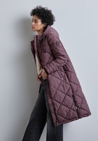 Lång, vadderad kappa i mörklila med huva, som har dragkedja och tryckknappsstängning samt diagonala sömmar. Bärs över grå jeans.