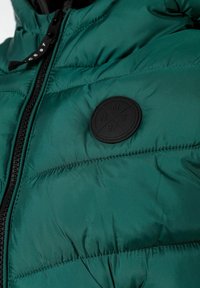 Zelena puffer jakna s kvadratnim uzorkom, prednjim zatvaračem i okruglim crnim grbom s natpisom "NMT 97" na prsima.