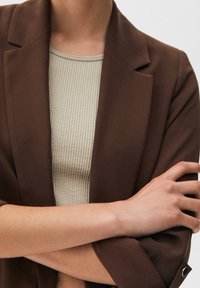 Brązowa marynarka o gładkiej fakturze, z wyraźnymi ramionami i klapami, zestawiona z jasnym, prążkowanym topem bez rękawów.