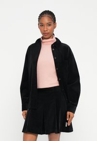 Črna kordna srajca na gumb preko svetlo roza turtlenecka, v kombinaciji s črno kordno krilom z naborki in majhnim logotipom.