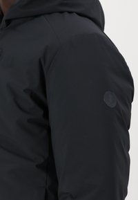 Veste noire isolée avec une finition lisse et mate. Elle est dotée d'une capuche et d'un écusson circulaire sur l'épaule gauche. Design sans coutures.