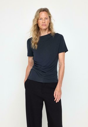 Donna con lunghi capelli biondi e mossi che indossa una camicia scura a maniche corte e pantaloni neri, in piedi con una mano in tasca su uno sfondo semplice.