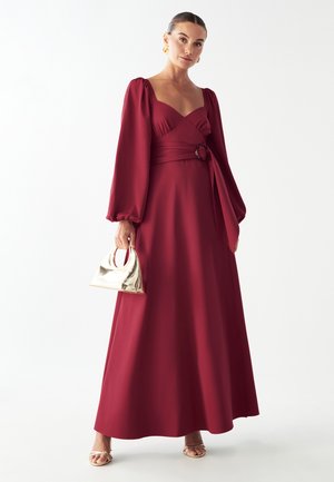 Largo vestido burdeos con mangas abullonadas y un corsé ajustado, que presenta una cintura ceñida con un acento circular. Combinado con un bolso metálico dorado.