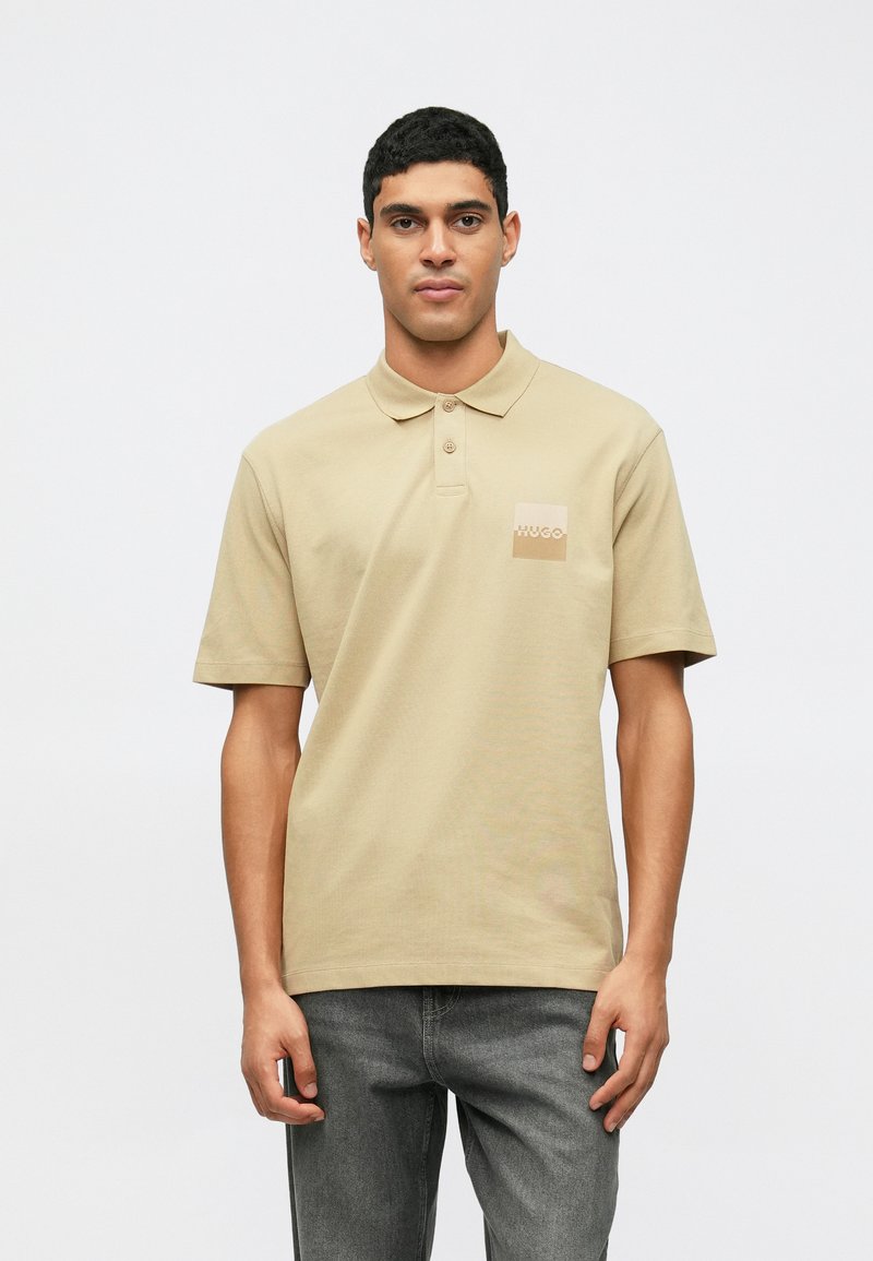 Homme portant un polo beige avec le logo "Hugo" sur la poitrine, associé à un jean gris foncé, se tenant devant un fond blanc uni.