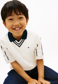 Χαμογελαστός νεαρός αγόρι με σκούρα μαλλιά, φοράει λευκό πόλο πουκάμισο με μπλε γιακά και ρίγες, καθισμένος με τα χέρια να ακουμπούν στα γόνατά του, σε λευκό φόντο.