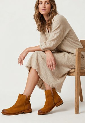 Kvinde med bølget hår sidder på en træstol iført en beige langærmet kjole og brune ankelstøvler med sidelås.