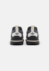 Sneaker grigio scuro con suole nere e dettagli crema. Presenta una parte superiore in rete testurizzata e un logo sulla linguetta del tallone. Lacci piatti e linguette di estrazione.