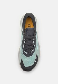Zapatilla de running sintética que presenta una parte superior de malla azul claro, acentos grises, puntera negra y una suela gris acolchada con patrones texturizados.