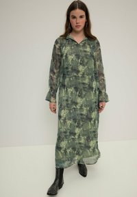 Largo vestido verde de patrón abstracto con cuello de camisa y mangas acampanadas, hecho de tela transparente, llevado con botas de tobillo negras con tachuelas.