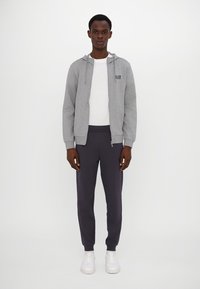 Siva zip-up dukserica s logom, bijela majica, tamnoplave trenirke i bijele tenisice. Glatka tkanina, ležeran kroj i minimalistički dizajn.