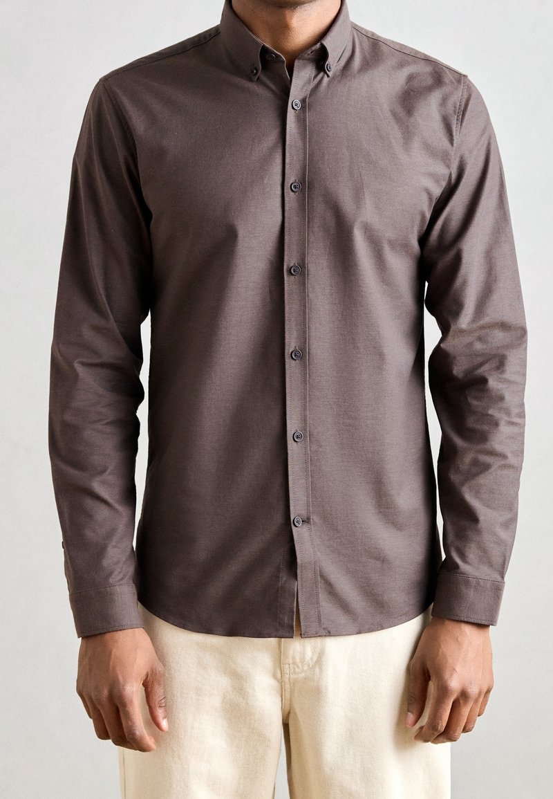 Homme portant une chemise marron ajustée à manches longues boutonnée et un pantalon beige clair, debout avec les bras détendus le long du corps.