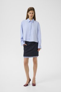 Donna in piedi con una camicia oversize azzurro chiaro con bottoni, una gonna a righe nere e pinstripe, e tacchi alti borgogna su uno sfondo bianco.