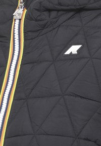 Schwarze Steppjacke mit diamantförmigem Steppmuster, ausgestattet mit einem zentralen Reißverschluss mit mehrfarbigen Streifen und einem weißen Logo-Patch auf der Brust.