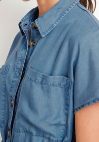 Světle modrá denimová košile s špičatým límečkem, krátkými rukávy, zapínáním na knoflíky vpředu a dvěma náprsními kapsami. Měkká textura a volný střih.