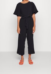 Mono negro con mangas cortas de vuelo y cintura anudada. La tela parece suave; cuenta con piernas anchas y cortadas. Se lleva con sandalias naranjas.