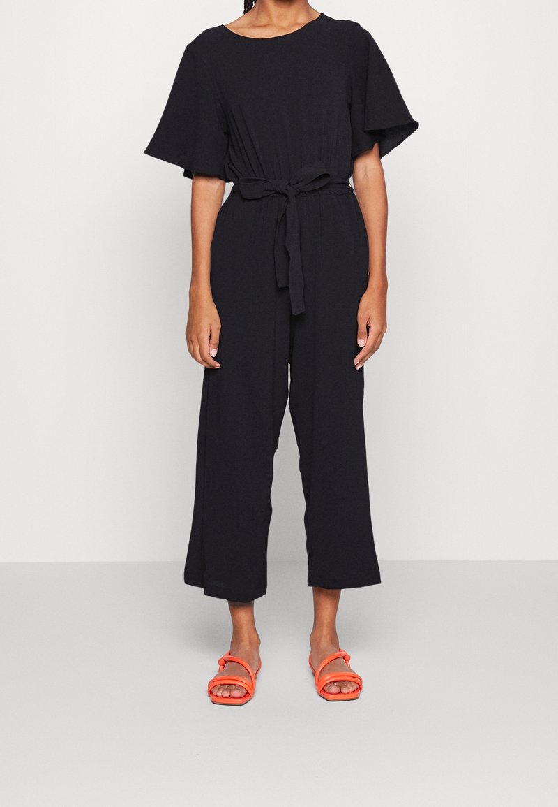 Mono negro con mangas cortas de vuelo y cintura anudada. La tela parece suave; cuenta con piernas anchas y cortadas. Se lleva con sandalias naranjas.