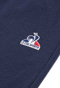 Marineblauwe stof heeft een wit geborduurd logo met rode en blauwe accenten, met een gestileerde haan en de tekst "LE COQ SPORTIF."