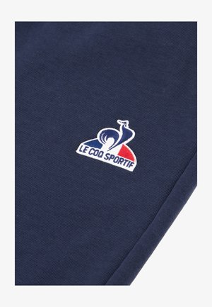 Un tejido azul marino presenta un logo bordado en blanco con acentos rojos y azules, mostrando un gallo estilizado y el texto "LE COQ SPORTIF."