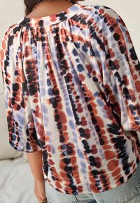 Femme portant un chemisier ample avec un col froncé et un motif tie-dye abstrait rouge, noir et bleu, associé à un jean bleu clair.