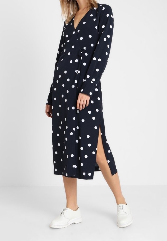 Robe portefeuille à pois navy avec des manches longues, détail de fente sur le côté et tissu léger, associée à des chaussures blanches.