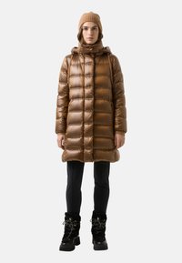 Braune Steppjacke mit vertikalem Steppmuster, Kapuze und figurbetonter Silhouette, kombiniert mit schwarzen Hosen und schwarzen Stiefeln mit Fellfutter.