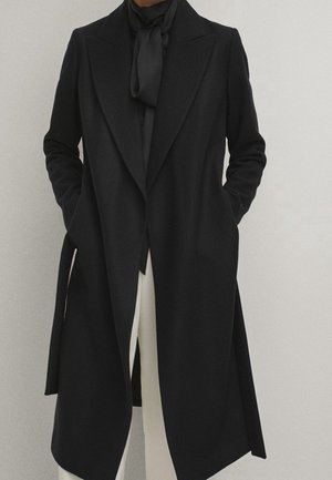 Personne portant un long manteau noir avec des revers, une écharpe noire et un pantalon blanc, se tenant devant un fond uni clair les mains dans les poches.
