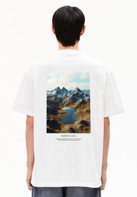 T-shirt blanc avec un imprimé graphique de montagnes et d'un lac au dos, avec le texte "Inspiré par la Nature" en dessous de l'image.