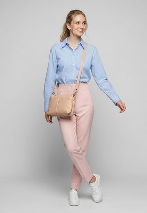 Ung kvinde iført en blå ternet skjorte, lyserøde bukser, hvide sneakers og en beige crossbody-taske, står i en afslappet stilling.