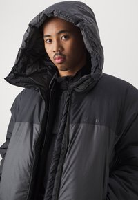 Puffer jacket in black and dark gray, featuring a large hood, zip closure, and quilted texture. Minimalist design with subtle branding.
