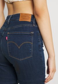 Mujer vistiendo jeans de mezclilla Levi's azul oscuro con costuras visibles en los bolsillos traseros y parche de cuero en la cintura.