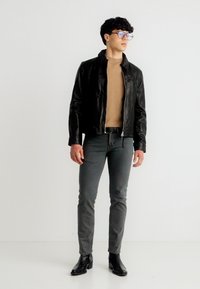 Νέος άνδρας με γυαλιά, μαύρο δερμάτινο μπουφάν, καφέ πουλόβερ, γκρι στενά παντελόνια και μαύρα μποτάκια στέκεται μπροστά σε λευκό φόντο.