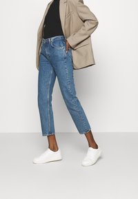 Vaqueros de mezclilla azul claro con cintura alta, con corte recto y dobladillo ligeramente deshilachado, combinados con una blusa negra y un blazer beige.