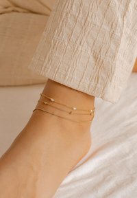 Złoty anklekt składający się z trzech delikatnych łańcuszków z małymi perłami i koralikami. Stopa spoczywa na jasnym, miękkim tle materiału.