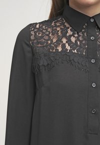 Maglietta nera con pannello in pizzo con design floreale sul petto, decorata con dettagli di frange e completata con bottoni neri.