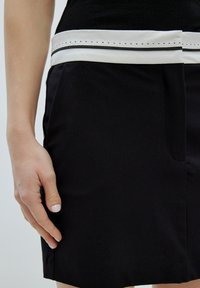 Zwarte rok met een witte band in de taille, met een gestikte detail. De stof lijkt glad en op maat gemaakt, met een zijzak.