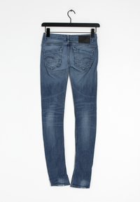 Siniset farkkulegginsit, joissa on sileä rakenne, viisi taskua ja hienovaraiset ompeluDetailit, roikkumassa mustassa ripustimessa valkoista taustaa vasten.