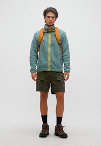 Νέος άνδρας που φοράει γαλάζιο σακάκι fleece, ελαι πράσινα cargo σορτς, ορειβατικά παπούτσια, μαύρες κάλτσες και πορτοκαλί σακίδιο πλάτης, στέκεται μπροστά από απλό φόντο.