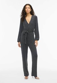 Juodas ir sidabrinis blizgus kombinezonas su ilgomis rankovėmis, giliais V formos iškirpte ir surišama juosmens dalimi. Audinys turi subtilų, tekstūruotą raštą.