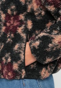 Fleece bunda s černým, růžovým a burgundským vzorem, s jemnou texturou a skrytou boční kapsou.