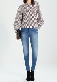 Raidallinen, pitkähihaisuus paita, jossa on puffihihat, yhdistetty vaaleansinisiin kapealöysiin farkkuihin ja mustiin nilkkureihin; laskokset ja kangasrakenne näkyvissä.