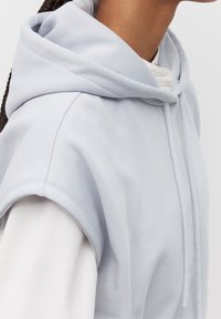 Světle modrá bezrukávová mikina s kapucí a stahovací šňůrkou, texturovaná látka a jemný povrch. Přes bílou dlouhou košili.