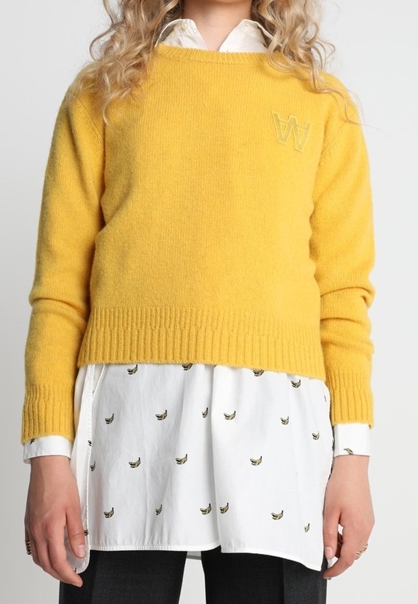 Pull jaune en tricot avec ourlet et poignets côtelés, orné d'un logo 'W' en relief. Superposé à une chemise blanche avec un petit imprimé banane.