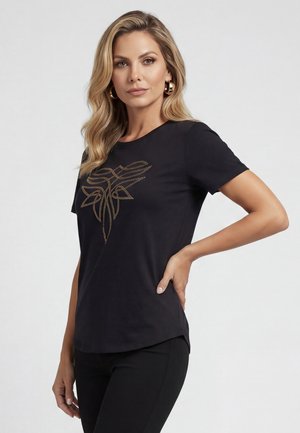 Kobieta o długich blond włosach, nosząca złote kolczyki koła i czarną koszulkę z złotym geometrycznym wzorem, pozująca z jedną ręką na biodrze.
