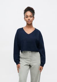 Pull en tricot bleu marine avec un col en V et des manches longues, associé à un pantalon taille haute gris clair. Les matériaux texturés sont visibles.